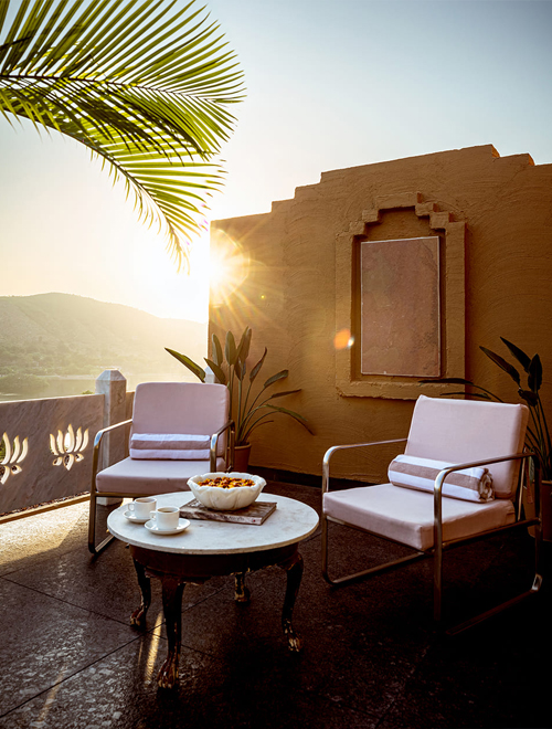 Experiences 6 Two chairs with cushions and a small round table with cups and a bowl are on a sunlit patio with a palm leaf and a stucco wall in the background.