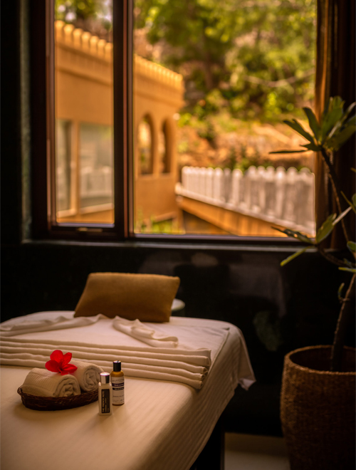 Experiences 1 A massage table with neatly folded towels, a red flower, and spa products is situated near a window with a scenic outdoor view.