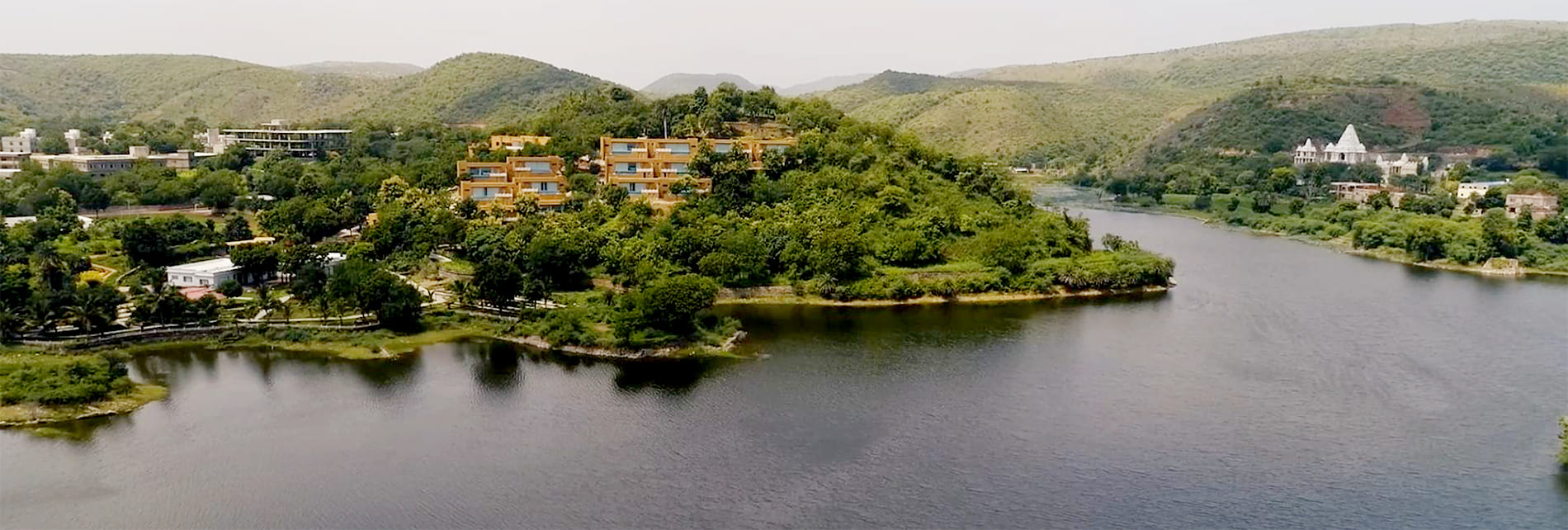 Contact us 1 Aerial view of a hilly terrain with green vegetation, a river winding through the landscape, and clusters of buildings including a white structure on the right side—an idyllic spot for a destination wedding.